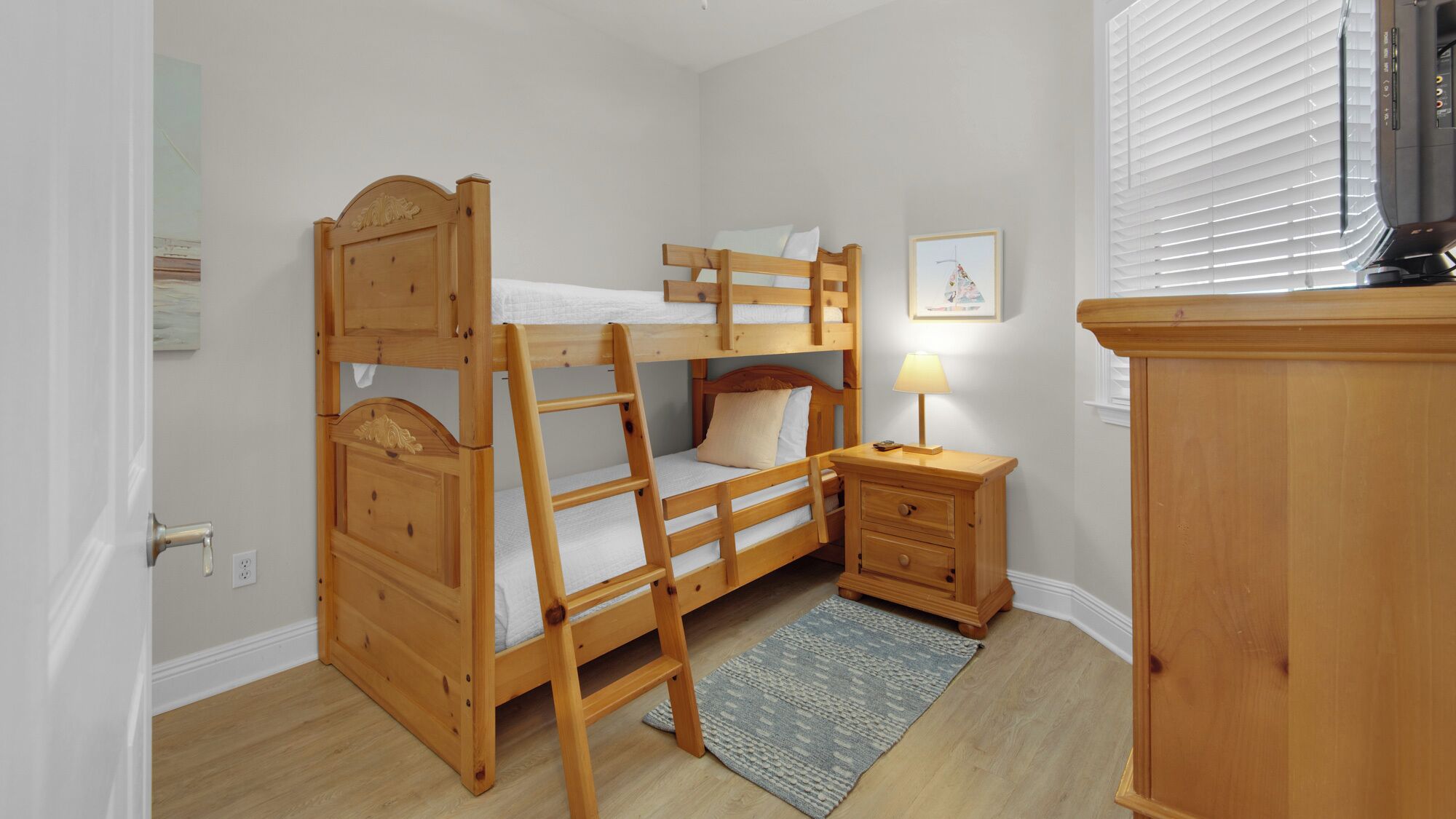 A bunk bed room with a wooden ladder, a bedside table with a lamp, a small cabinet, and a framed picture on the wall.