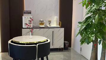 A modern hotel room with a marble floor, a round dining table with chairs, a minibar, and a potted plant.