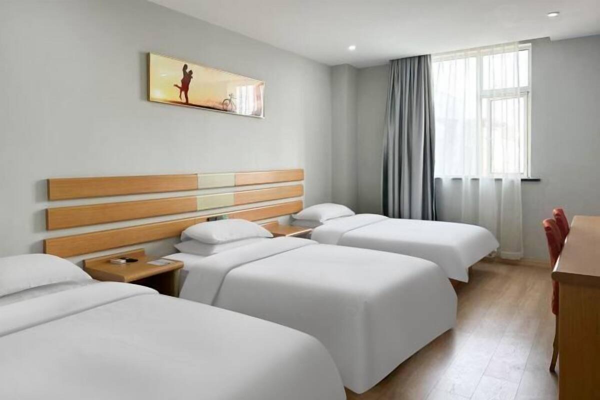 Habitación de hotel con tres camas, cabecera de madera, un cuadro en la pared, ventana con cortinas y un escritorio.