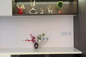 Une cuisine avec des armoires blanches, un plan de travail blanc et une étagère ornée d’objets décoratifs, notamment des fleurs et un petit arbre.