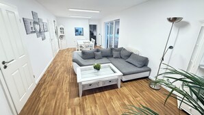 A modern living room with a grey sectional sofa, a white coffee table, and a floor lamp. There is a dining area with a white table and chairs in the background.
