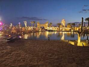 Un resort nocturno con una piscina grande, palmeras y estructuras iluminadas.