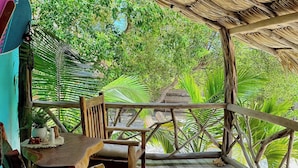 Une terrasse extérieure en bois, dotée d’un toit de chaume, de chaises en bois et d’une table avec un coussin.