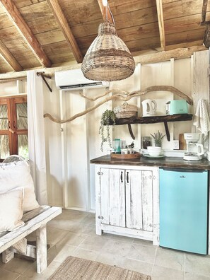 Una cocina rústica con techo de madera, una lámpara colgante de mimbre, un armario blanco con refrigerador azul y un banco de madera.