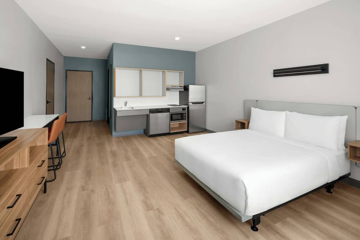 A hotel room with a bed, a desk, a chair, a TV, a kitchenette, and a bathroom.