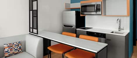 A compact kitchen with a microwave, refrigerator, and dishwasher. There are two orange bar stools and a gray sofa with a patterned pillow.
