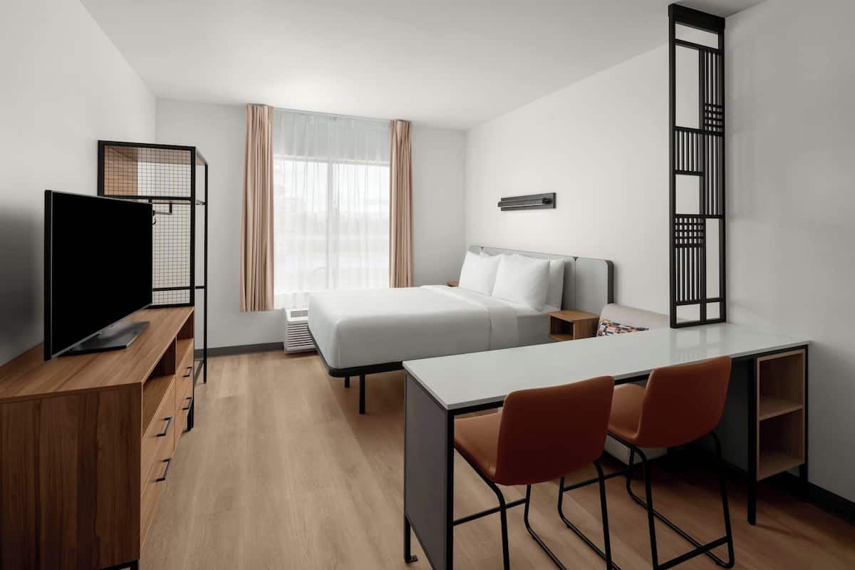 A modern hotel room with a bed, a desk, two chairs, a TV, and a window with curtains.