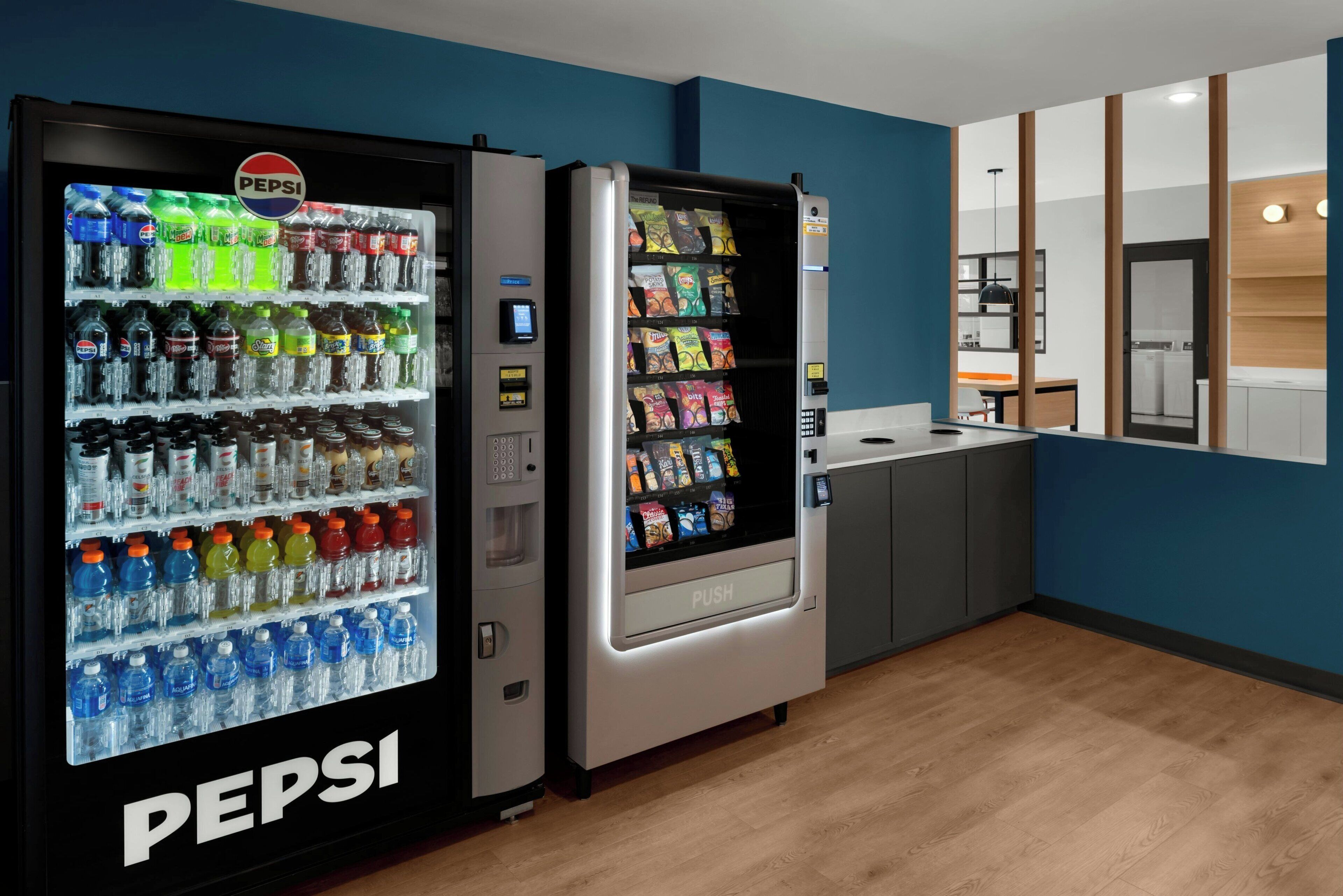 Un espace comprenant un distributeur automatique de boissons, avec des produits Pepsi et d’autres boissons, un autre distributeur à bouton-poussoir, ainsi qu’un coin cuisine équipé de placards et d’un comptoir.