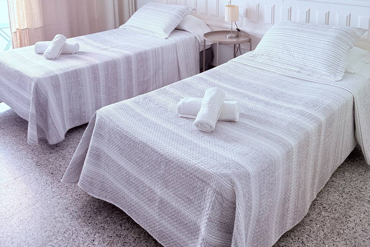 A hotel room with two beds, a small bedside table with a lamp, an air conditioning unit, and a window with curtains.
