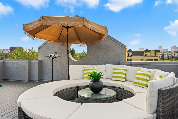 Een terras op het dak met een gebogen witte bank, een ronde glazen tafel en een grote beige parasol.
