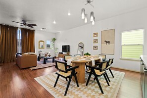 A modern living room with a dining area, wooden flooring, a ceiling fan, and a large window with blinds.