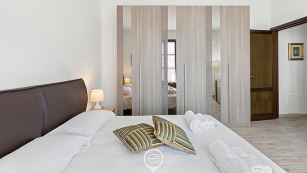 Una habitación de hotel moderna con una cama grande, dos almohadones decorativos, una mesita de noche con lámpara y vistas a un baño a través de una puerta corredera.