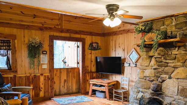 A cozy cabin interior with wooden walls, a stone fireplace, a television, and a wooden sofa. - Once a Blacksmith shop, now a 3-bedroom cabin beauty in the heart of the Ozarks (Yellville)