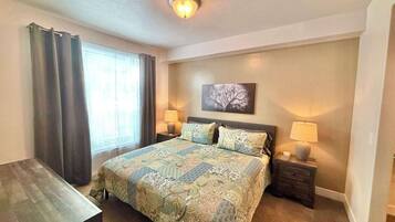 A hotel room with a large bed, two bedside tables with lamps, a desk, a window with curtains, and a ceiling light.