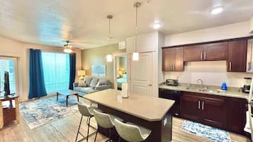 A modern kitchen with dark wooden cabinets, a central island with bar stools, a dining area with a table and chairs, and a living room with a sofa and a TV.