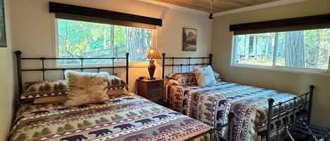 A bedroom with two beds, wooden ceiling, and windows with a view of trees.