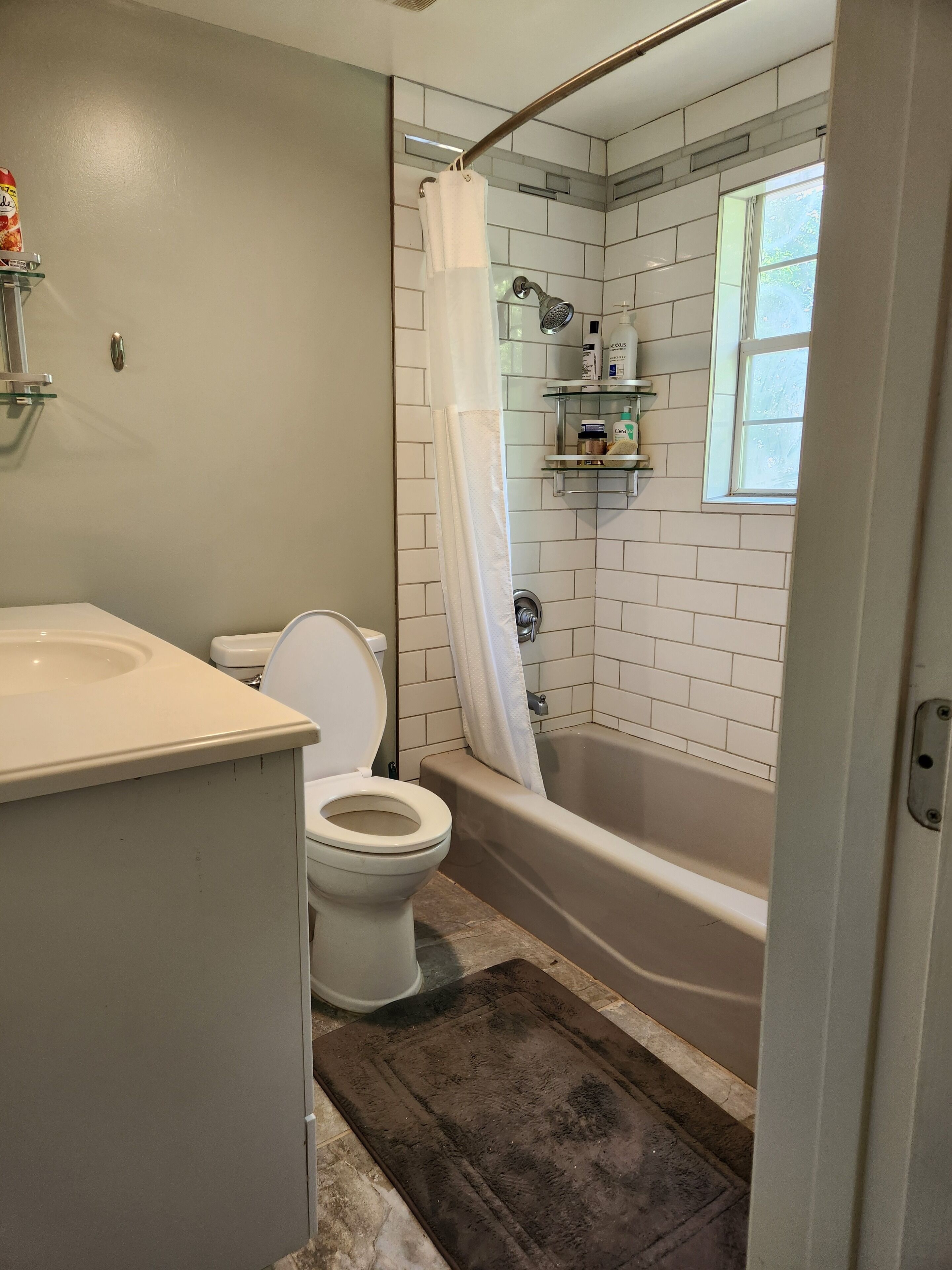 A bathroom with a toilet, sink, and bathtub. There is a shower curtain, a window, and a shelf with toiletries.