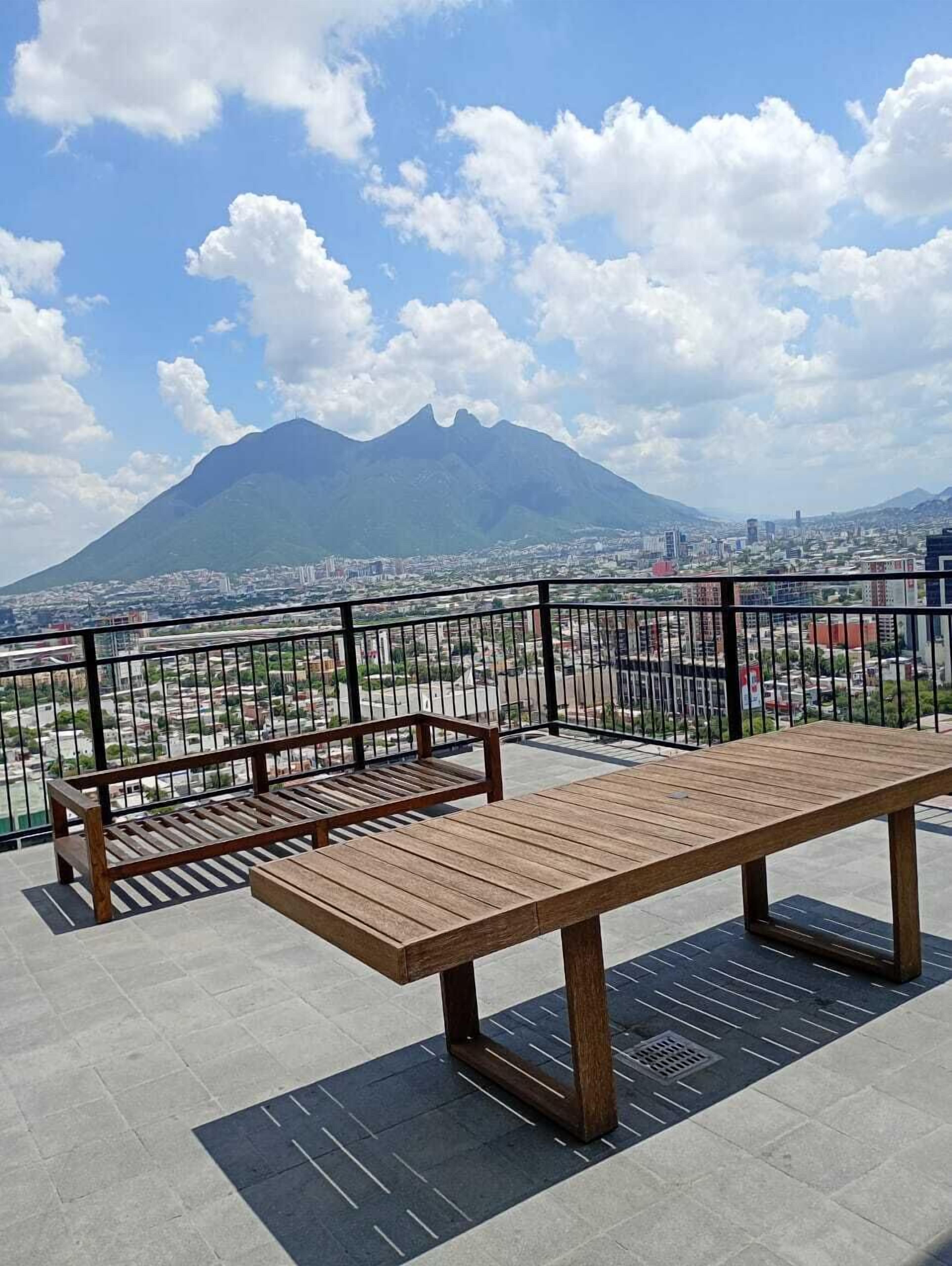 Un banco de madera con barandilla de metal sobre una plataforma de baldosas, con vistas a un paisaje urbano y una montaña.