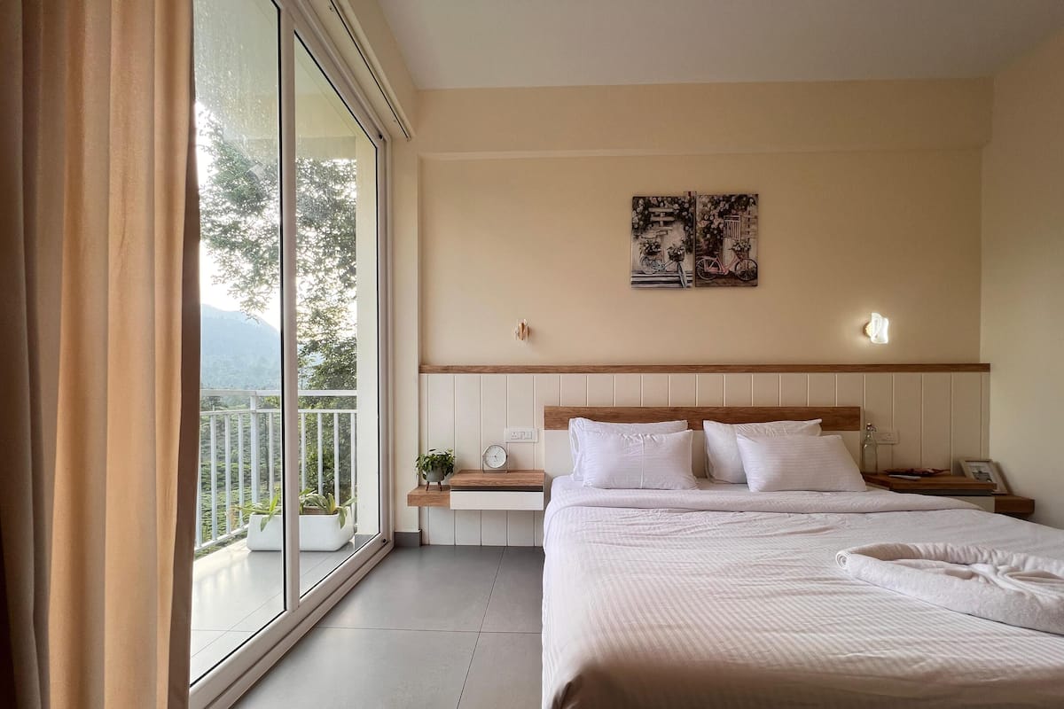 A hotel room with a bed, bedside tables, a nightstand, a wall-mounted clock, a small potted plant, and a painting.