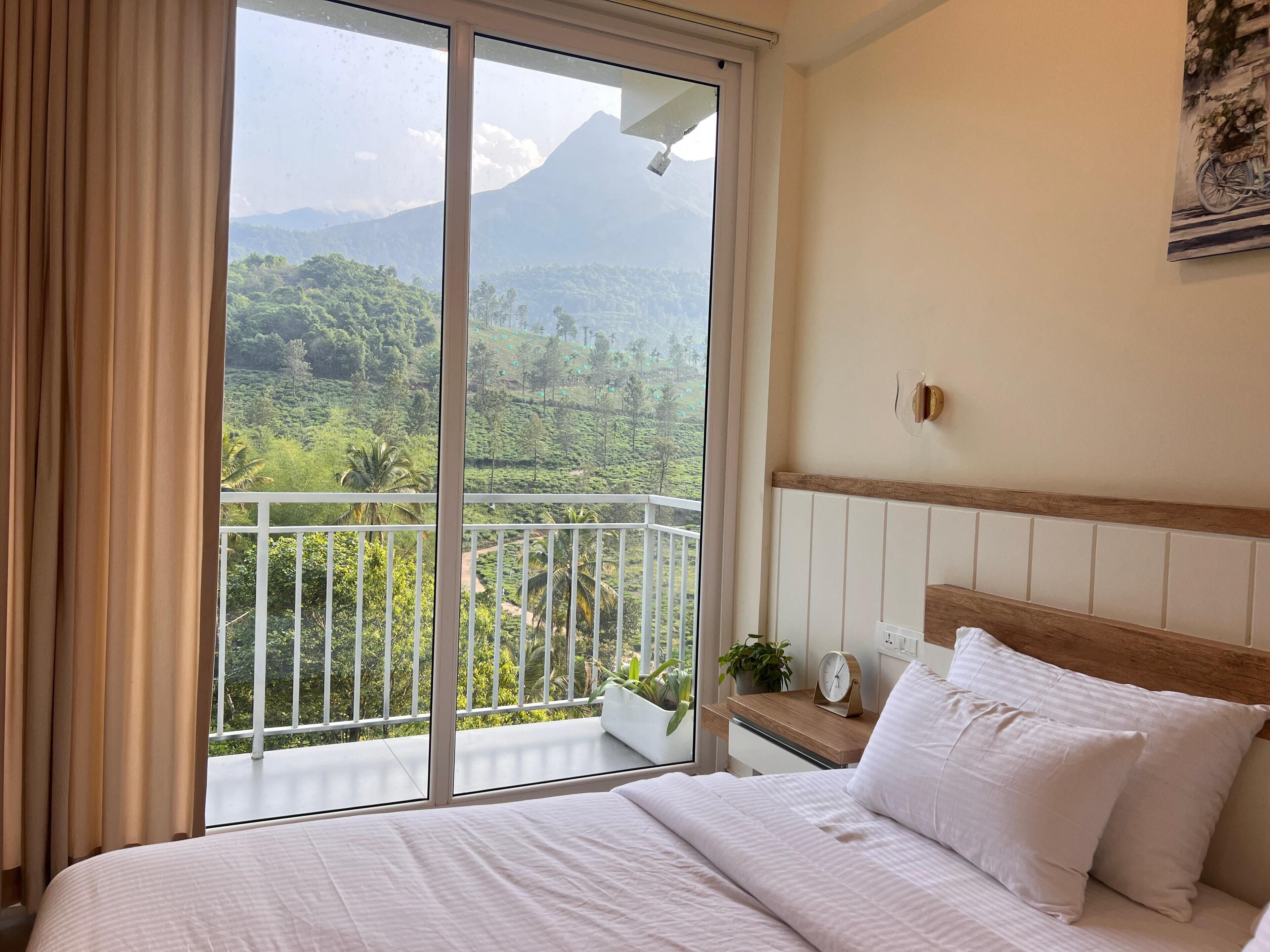 A hotel room with a bed, a nightstand with a clock, a potted plant, and a view of a lush landscape with a mountain.