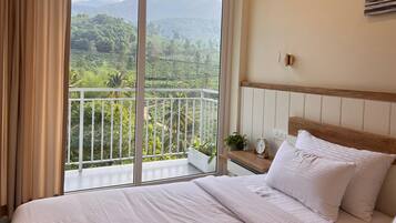 A hotel room with a bed, a nightstand with a clock, a potted plant, and a view of a lush landscape with a mountain.