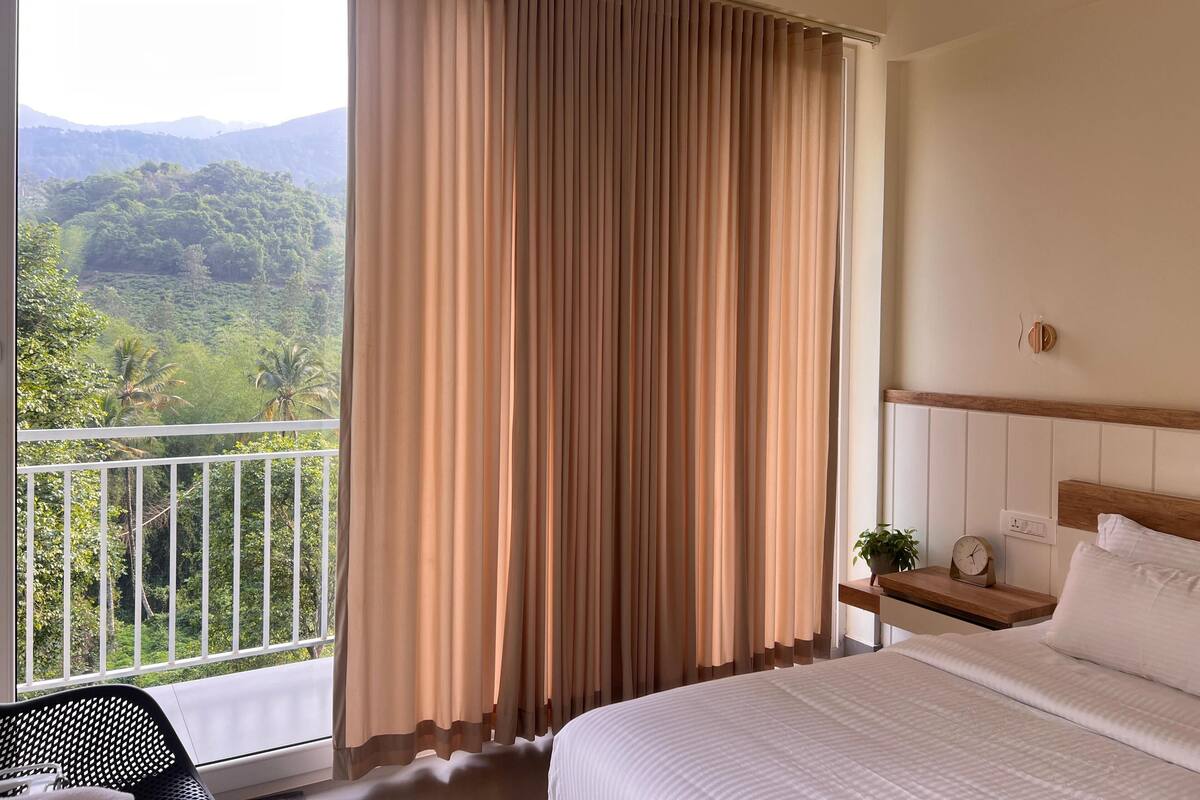 A hotel room with a bed, a nightstand, a clock, and a view of a lush landscape through large curtains.