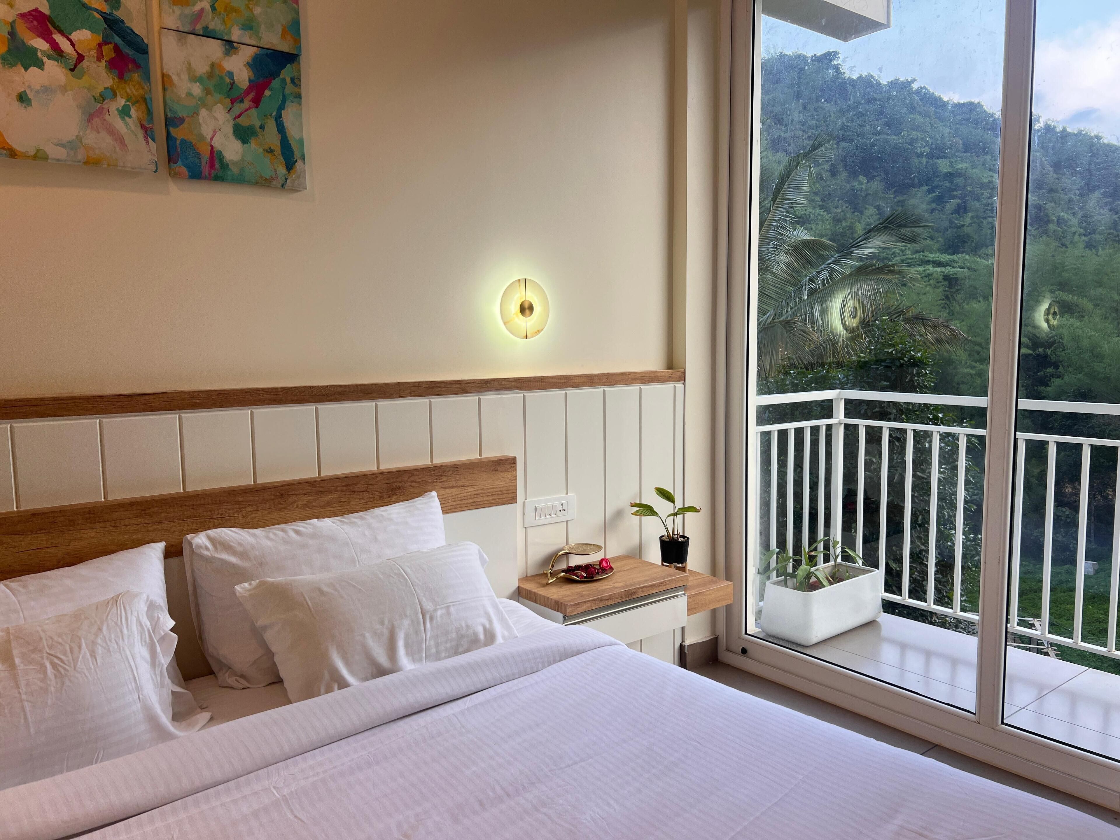 A hotel room with a large bed, a nightstand, a wall-mounted light, and a balcony with a view of greenery.