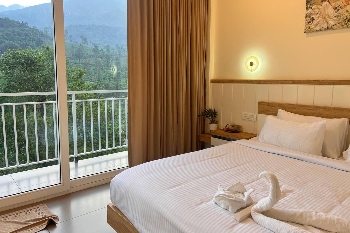 A hotel room with a large bed, a bedside table with a plant, a painting on the wall, and a balcony with a mountain view.