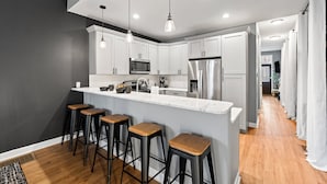 A modern kitchen with white cabinets, a stainless steel refrigerator, a microwave, and a large island with stools. - The Journey Home | JZ Vacation Rentals (St. Louis)