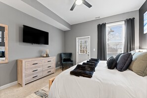A bedroom with a large bed, a TV mounted on the wall, a wooden dresser, a chair, and a window with curtains. - The Journey Home | JZ Vacation Rentals (St. Louis)