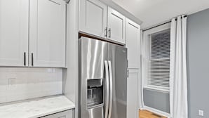 A modern kitchen with white cabinets, a stainless steel refrigerator, and a window with blinds. - The Journey Home | JZ Vacation Rentals (St. Louis)