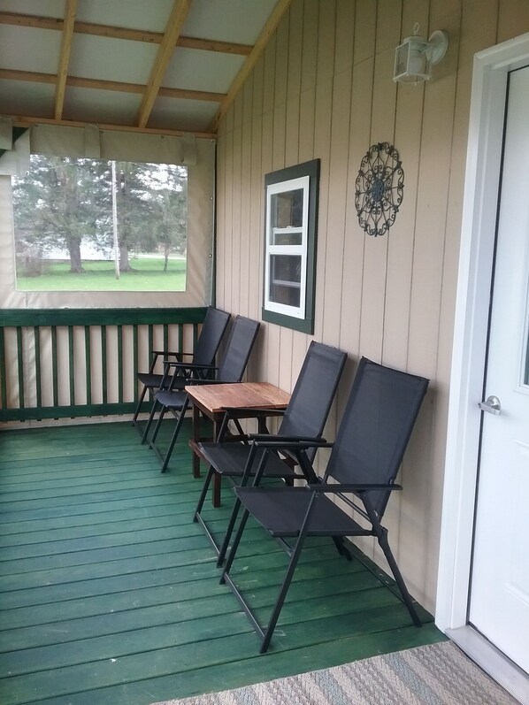 A covered porch with four black chairs and a wooden table, a white door, and a window with a view of trees. - River Bungalow in lovely Dunn's Eddy, Youngsville with AC, WiFi & Fire ring. (Youngsville)