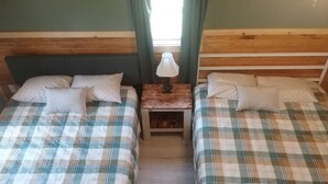 Een kamer met twee bedden, elk voorzien van een geruite sprei en kussens, een klein houten nachtkastje met een lamp, en een raam met gordijnen.