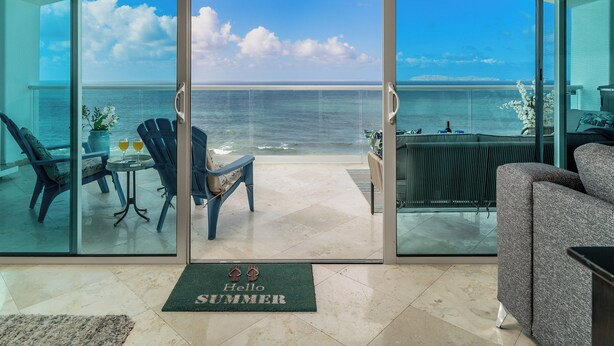 Un balcon avec vue sur l’océan, une chaise, une table avec des boissons et un paillasson sur lequel est écrit « Bonjour, l’été ! ».