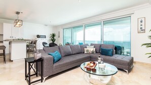 Un salon moderne avec un canapé d’angle, une table basse en verre et une vue sur l’océan grâce à de grandes fenêtres.