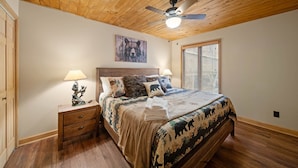 A bedroom with a wooden ceiling, a bed with a forest-themed bedspread, a headboard with a bear painting, a bedside table with a lamp, and a window with blinds. - Spacious 6BR mountain retreat with hot tub, game room, and views. (Beech Mountain)