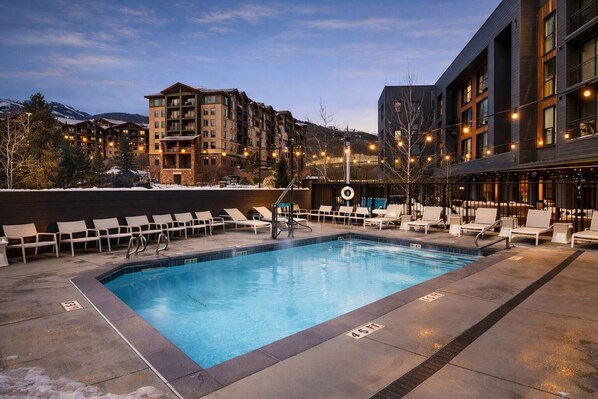 Un área de piscina con sillones de descanso y una piscina, rodeada de edificios con balcones e iluminación.