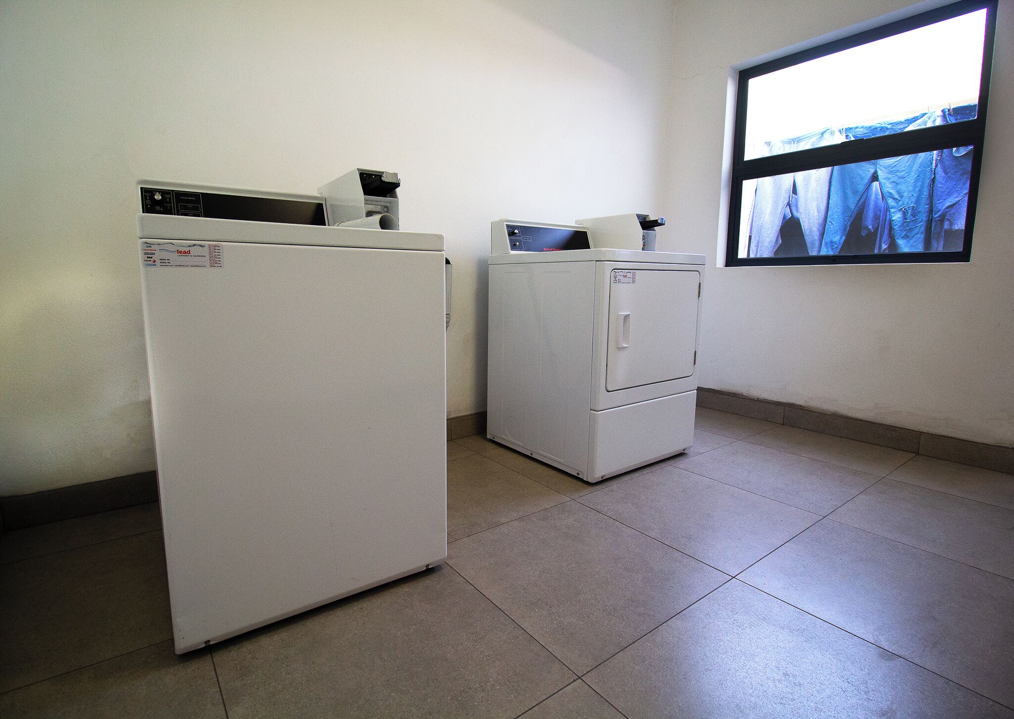 A laundry room with two white appliances, one larger and one smaller, and a window with a view of clothes hanging outside.