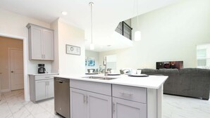 A modern kitchen with grey cabinets, a dishwasher, and a dining area with a grey sofa. - Champions Gate Gem – Pool, Spa, Near Parks (Davenport)