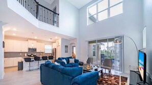 Un vaste espace de vie comprenant un canapé d’angle bleu, une table à manger avec des chaises, une cuisine en arrière-plan et un balcon avec un téléviseur.