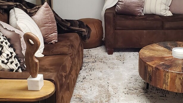 Ein Wohnzimmer mit zwei braunen Sofas, einem runden Holztisch, einem weißen Teppich und einer großen Grünpflanze.