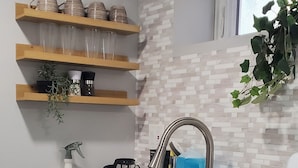 Eine moderne Küche mit weißen Schränken, einer doppelten Spüle und Edelstahlgeräten. Darüber befinden sich Holzregale mit Gläsern und einer Pflanze.
