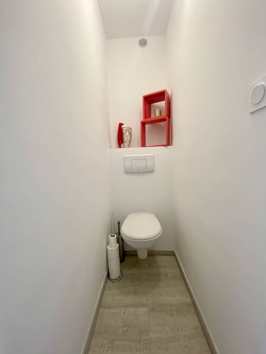 A narrow bathroom with a white toilet, a red shelf with a bottle, and a white toilet paper holder.