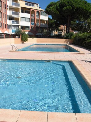 Ein Schwimmbecken mit gefliestem Sonnendeck, umgeben von einem Gebäude mit Balkonen und einem Baum.
