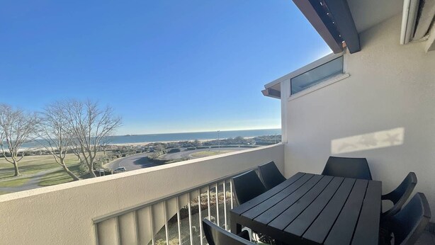 A balcony with a table and chairs, overlooking a coastal road and ocean.