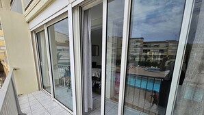 A balcony with sliding glass doors, a tiled floor, and a view of a swimming pool and buildings in the background. - CAP D'AGDE / Le Môle : Réf 0073 (CAP D'AGDE)