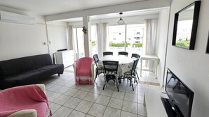 A living room with a black sofa, a dining table with chairs, a TV, and a window with curtains. - CAP D'AGDE / Le Môle : Réf 0073 (CAP D'AGDE)