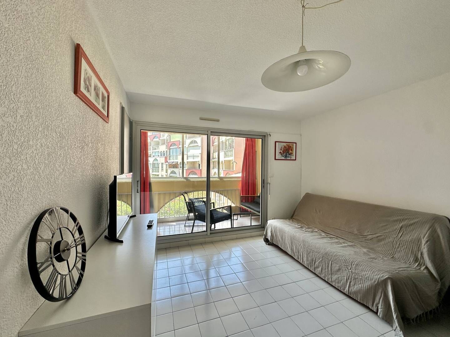 A room with a bed, a clock, a TV, and a balcony with a view of buildings.