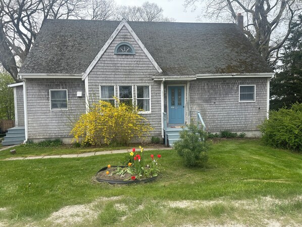 A two-story house with a gabled roof, a front porch, and a small garden with flowers. - Sawyer Cove Cottage (Jonesport)