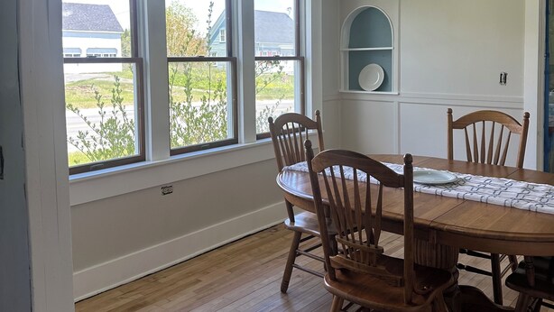 A dining area with a wooden table and chairs, large windows, a hanging light fixture, and a white wall with a shelf.